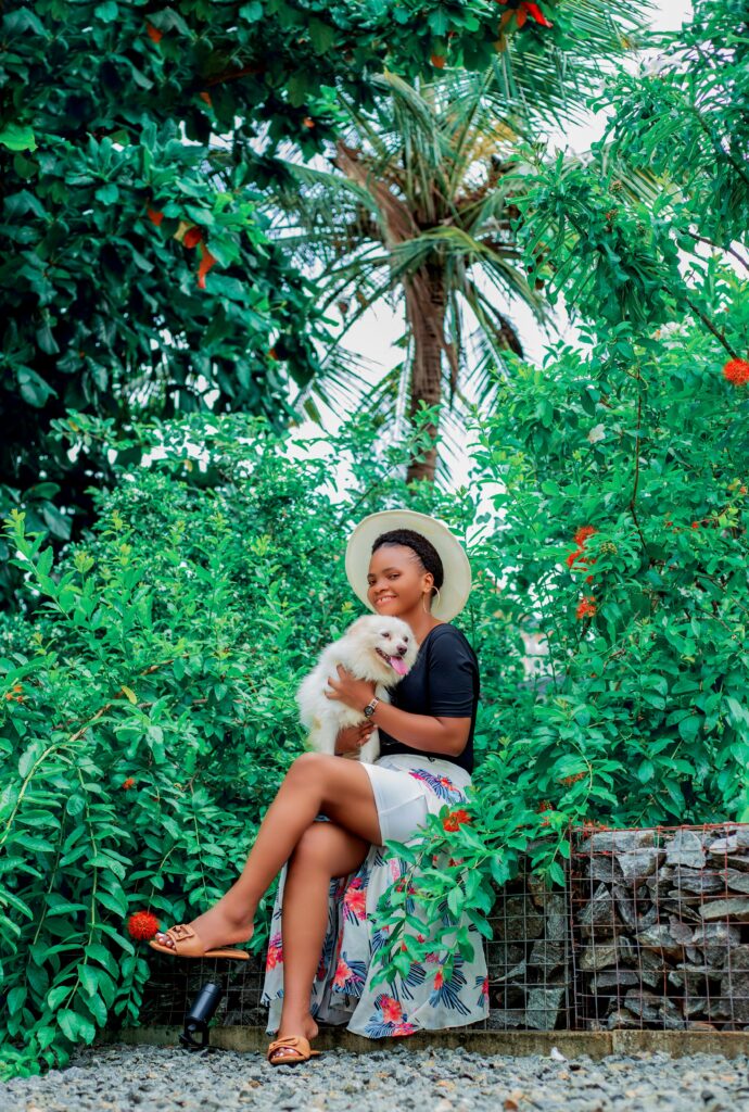pexels-photo-29557488-29557488 Smiling woman sitting amidst lush green foliage, holding her fluffy dog and wearing a sun hat.